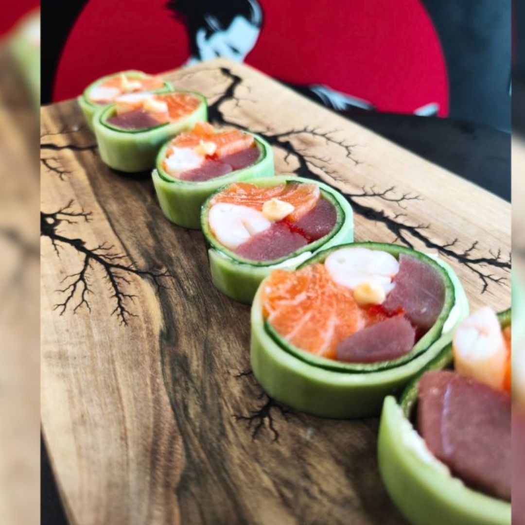 Nowoczesne sushi: rolki z ogórka wypełnione łososiem, tuńczykiem i krewetkami, ułożone na drewnianej desce z motywem drzewa, w tle czerwony okrąg z sylwetką.