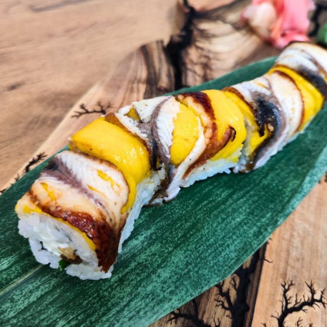 Sushi roll z węgorzem i mango na zielonym liściu, ułożone na drewnianej desce z wypalonym wzorem drzewa.