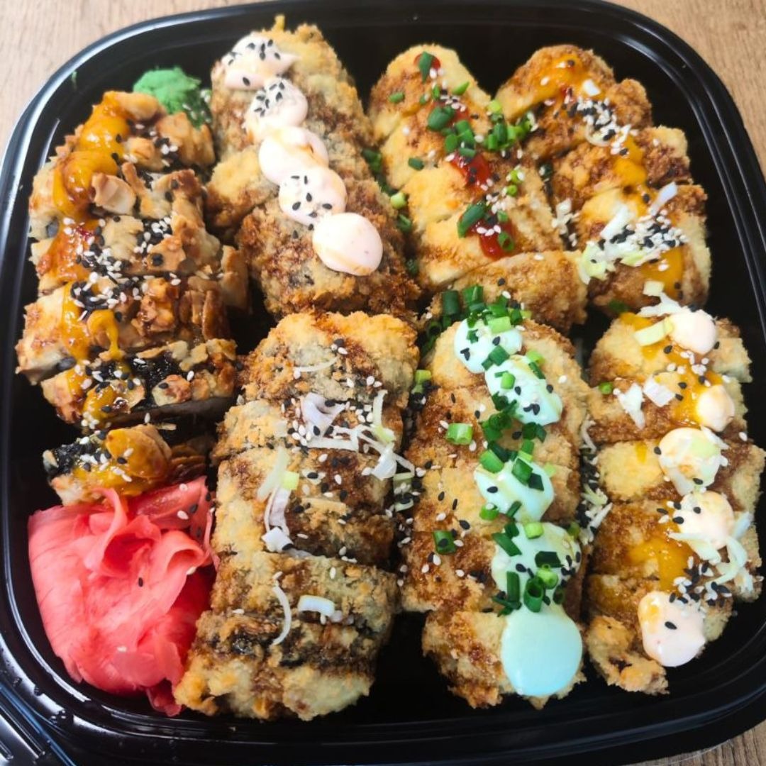 Różnorodne rolki sushi tempura w czarnym pojemniku, udekorowane sezamem, sosem i szczypiorkiem, z imbirem na boku.