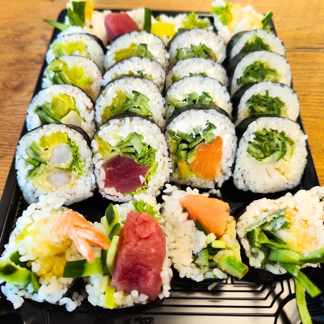 Zestaw różnorodnych rolek sushi na czarnej tacy, z widocznymi składnikami: tuńczyk, łosoś, krewetka w tempurze, ogórek i warzywa liściaste, ułożonych na drewnianym stole.