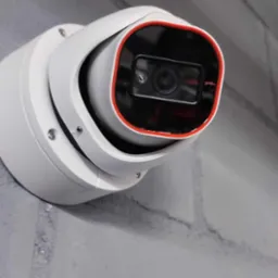 Biała kamera monitoringu kopułkowa z czerwoną obwódką, zamontowana na szarej ścianie z cegieł.