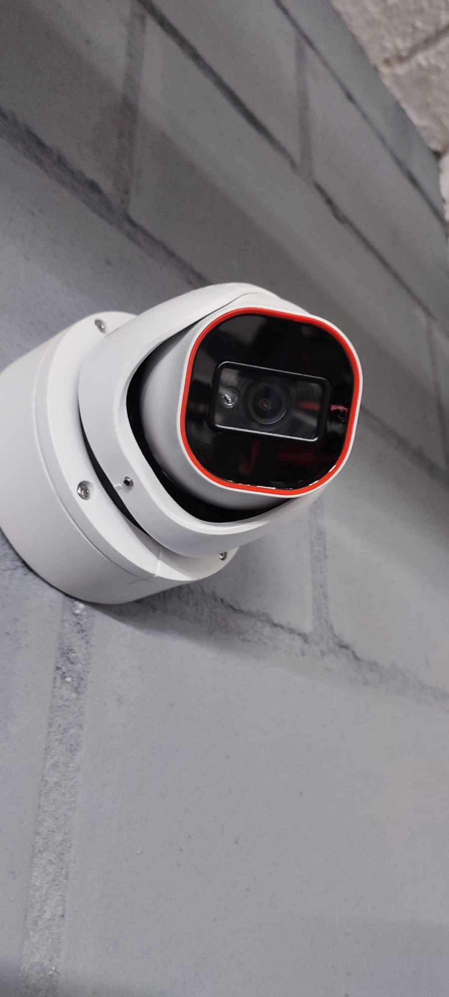 Biała kamera monitoringu kopułkowa z czerwoną obwódką, zamontowana na szarej ścianie z cegieł.