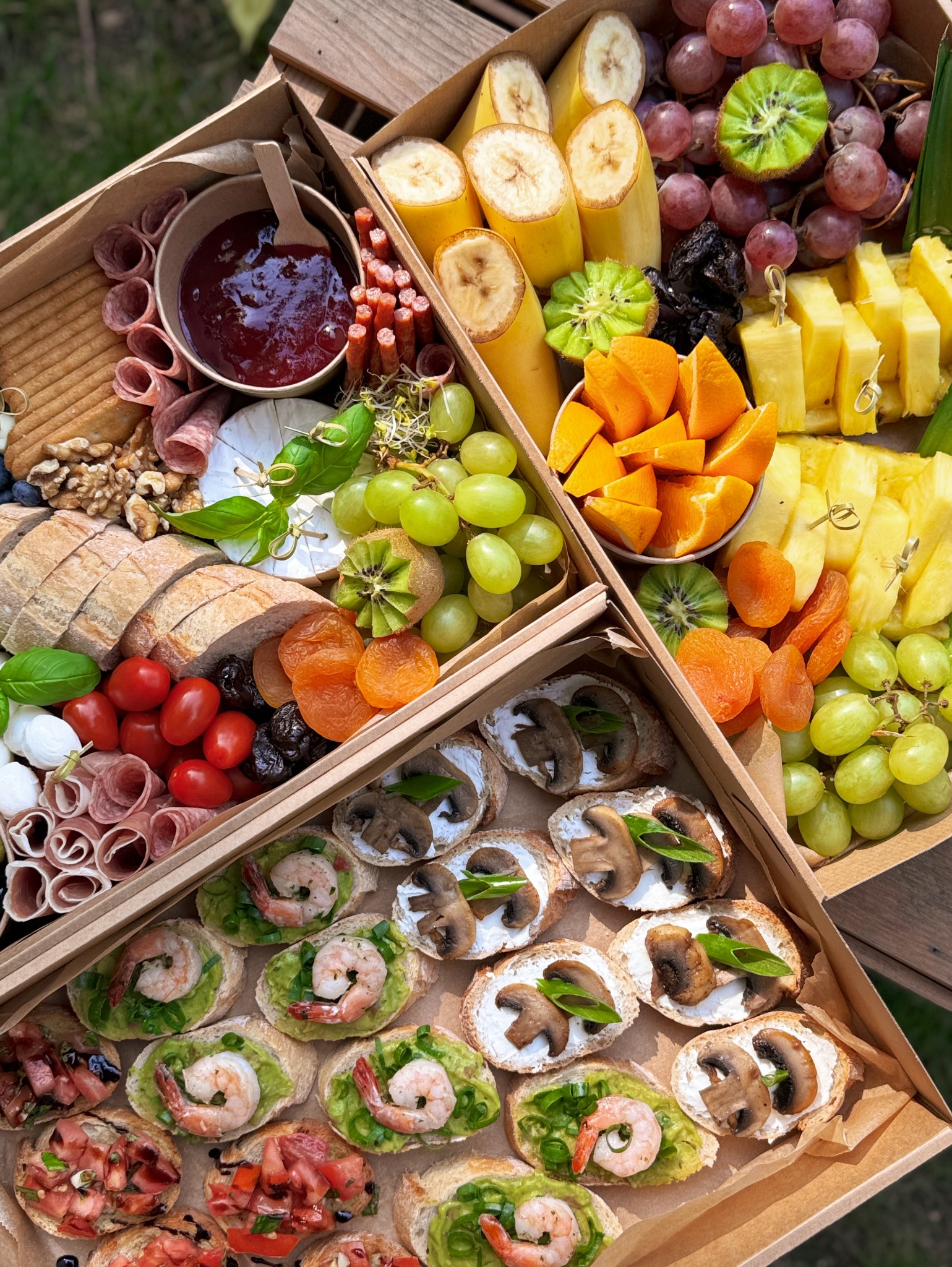 Elegancki catering w pudełku: kanapki z krewetkami i awokado, grzanki z grzybami, owoce, sery, wędliny i dżem w tekturowym pudełku z przegródkami na drewnianym stole.