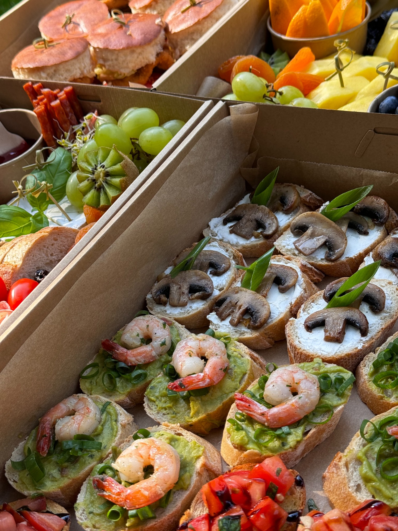 Różnorodna selekcja kanapek i przekąsek w kartonowym pudełku, w tym mini burgery, grzanki z awokado i krewetkami, grzanki z pieczarkami i twarożkiem, oraz owoce takie jak kiwi i winogrona.