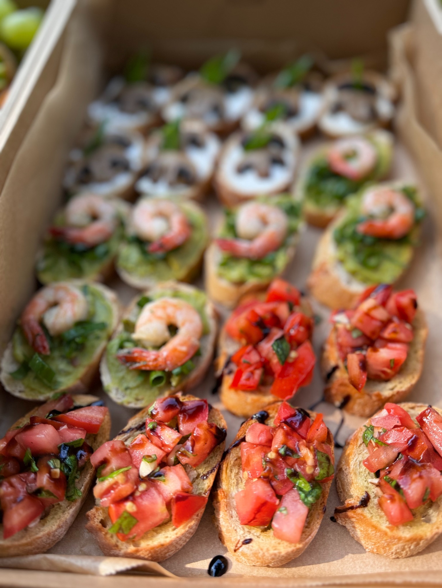 Różnorodne kanapki na pieczywie, w kartonowym pudełku: z krewetkami i zielonym pesto, z pomidorami i bazylią skropione octem balsamicznym oraz z pieczarkami.