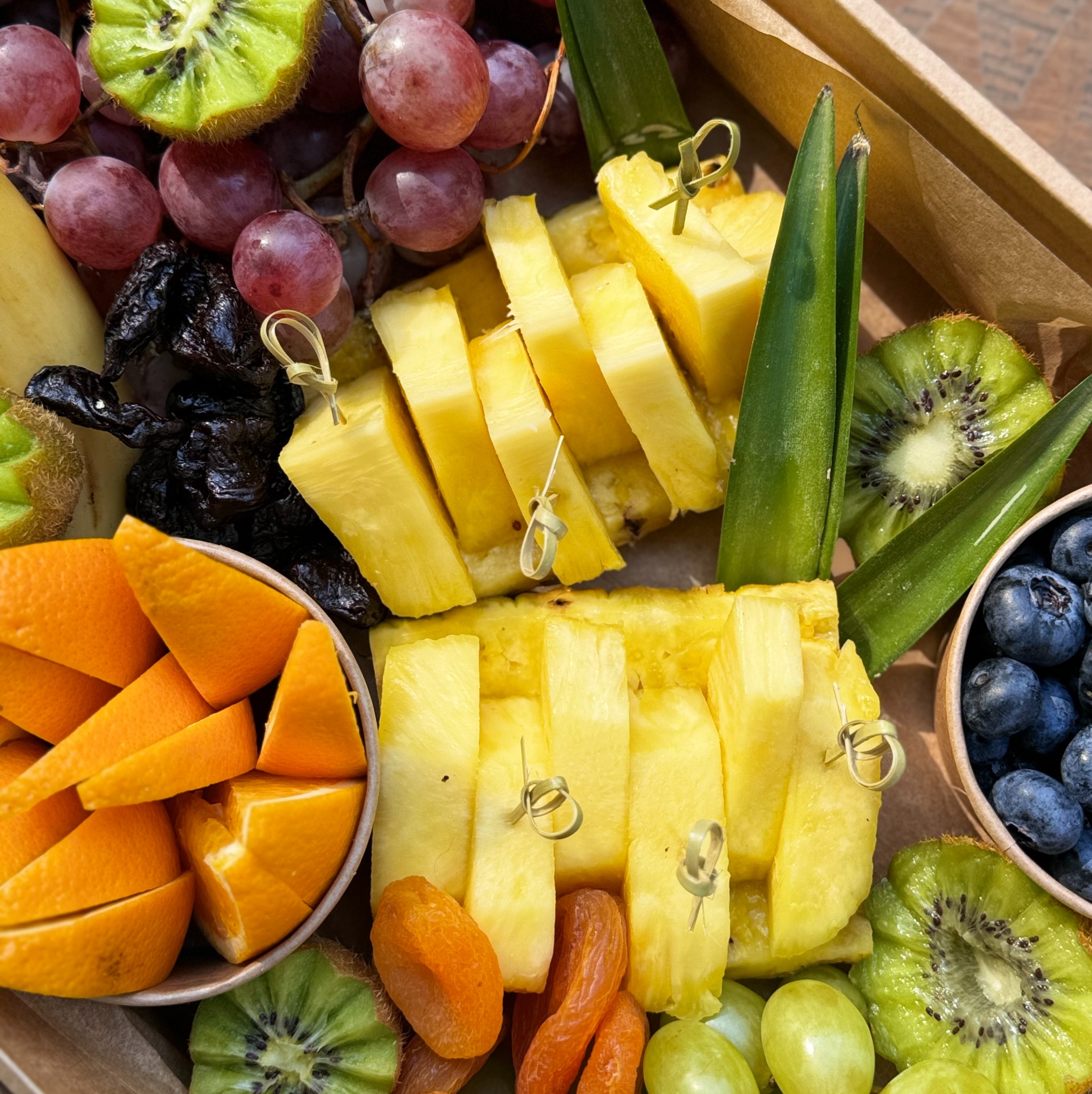 Dekoracyjna kompozycja z owoców: ananasa w kostkach z bambusowymi wykałaczkami, pomarańczy, kiwi, ciemnych śliwek suszonych, winogron i borówek w kartonowym pudełku.