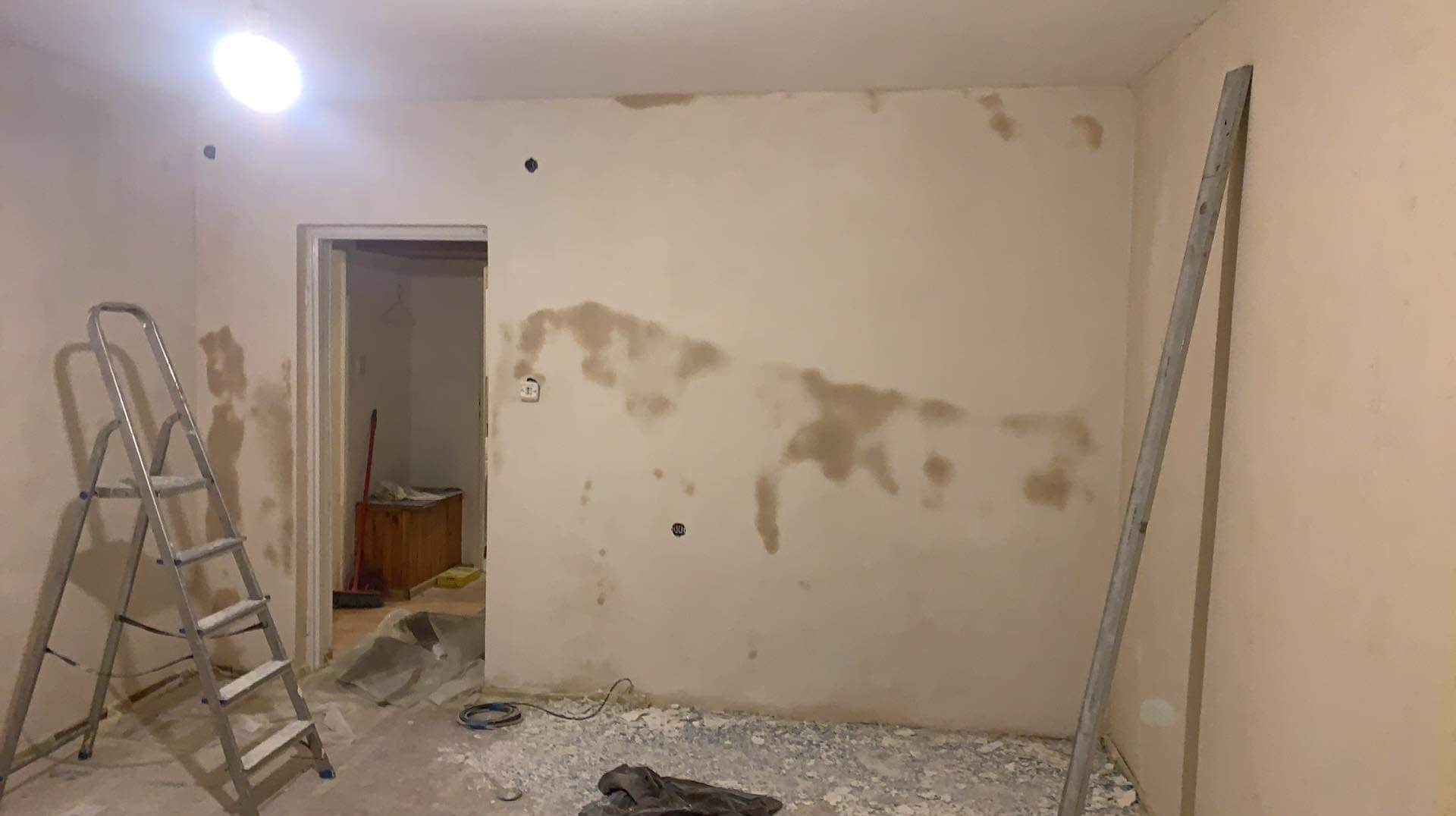 Przygotowanie ściany do malowania: widoczne plamy po zagruntowaniu, drabina malarska, listwa przypodłogowa, resztki gruzu na podłodze, gniazdko elektryczne.