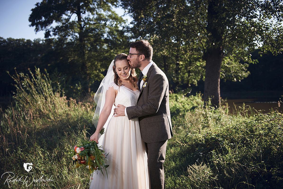 Para młoda w plenerze: Panna młoda w koronkowej sukni i welonie trzyma bukiet, pan młody w tweedowym garniturze całuje ją w czoło na tle zieleni.