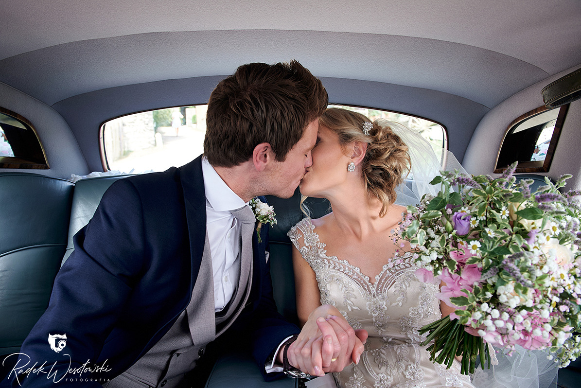 Pocałunek młodej pary w samochodzie w Warzymicach, panna młoda trzyma bukiet z fioletowymi akcentami, widoczny pierścionek na palcu.