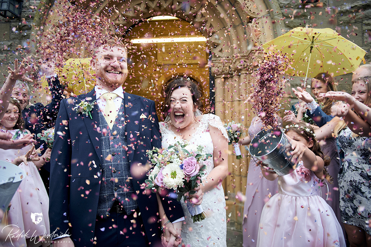 Uśmiechnięta para młoda wychodząca z kościoła, obsypywana konfetti w otoczeniu gości, dziewczynka z wiadrem konfetti na pierwszym planie.