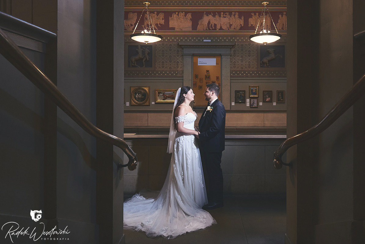 Para młoda w eleganckich strojach pozuje w muzeum sztuki, otoczona obrazami i dekoracyjnymi fryzami.