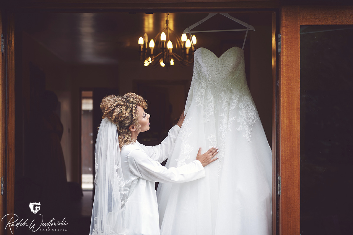 Panna młoda w białym szlafroku i welonie podziwia wiszącą na wieszaku suknię ślubną z koronkowymi zdobieniami, widoczną w otwartych drzwiach, w tle żyrandol.
