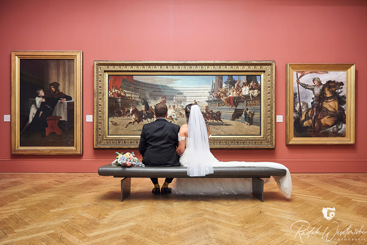 Para młoda siedząca na ławce w muzeum, oglądająca obrazy w złotych ramach, bukiet ślubny leży obok nich, parkiet na podłodze.