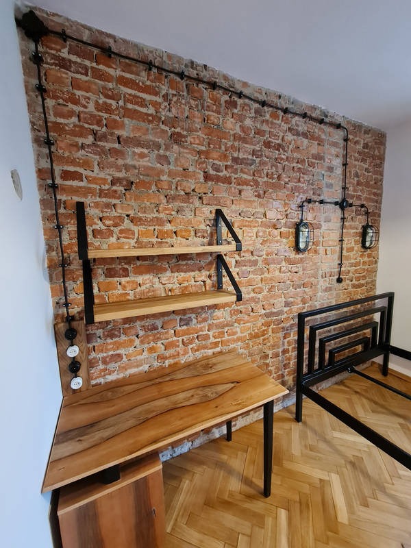 Loftowe wnętrze z odsłoniętą cegłą: instalacja elektryczna natynkowa z włącznikami, drewniany blat biurka i półki, metalowa rama łóżka, parkiet w jodełkę.