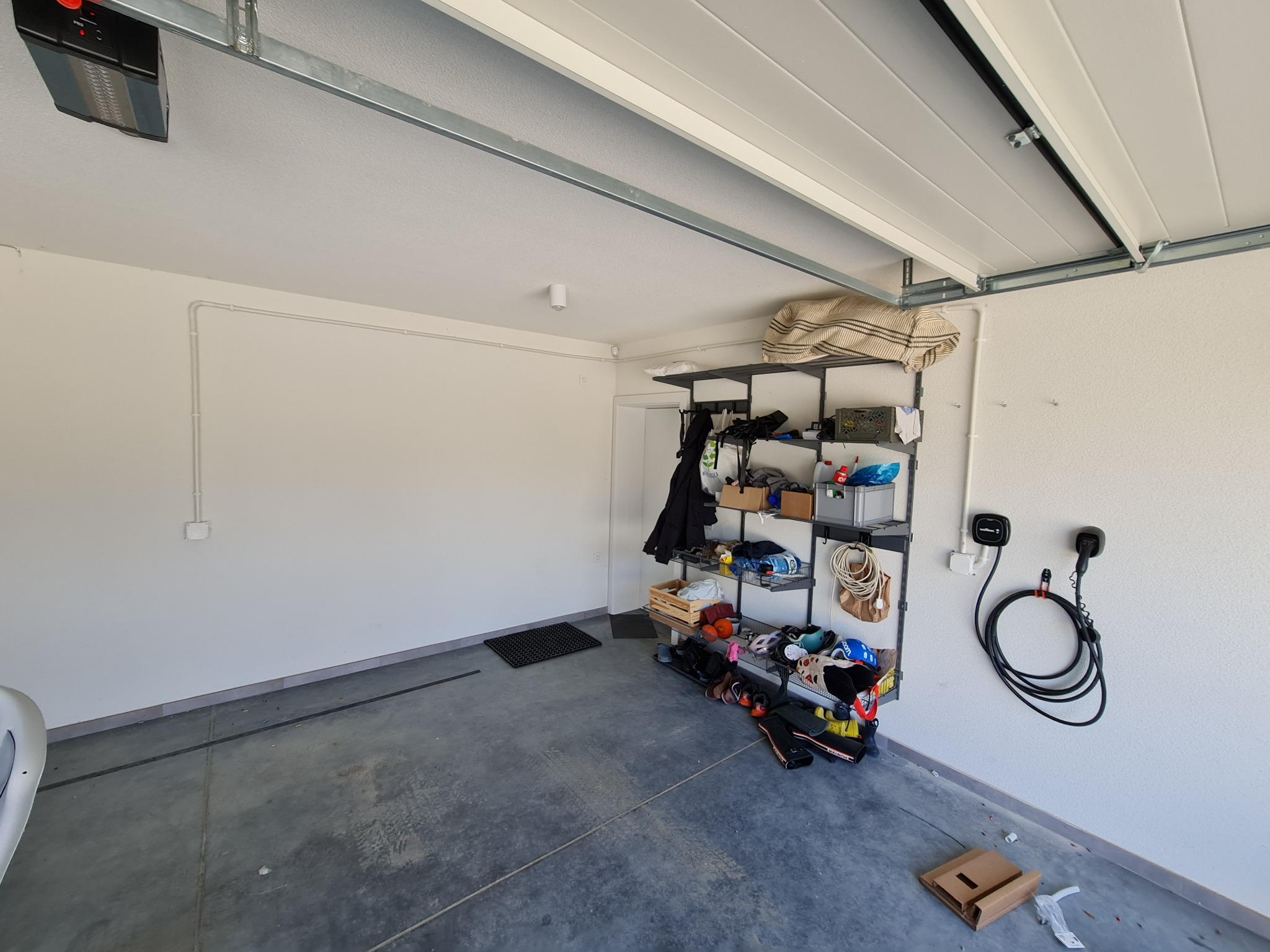 Garaż z zamontowaną ładowarką do samochodu elektrycznego na ścianie, widoczne przewody elektryczne i automat do bramy garażowej.