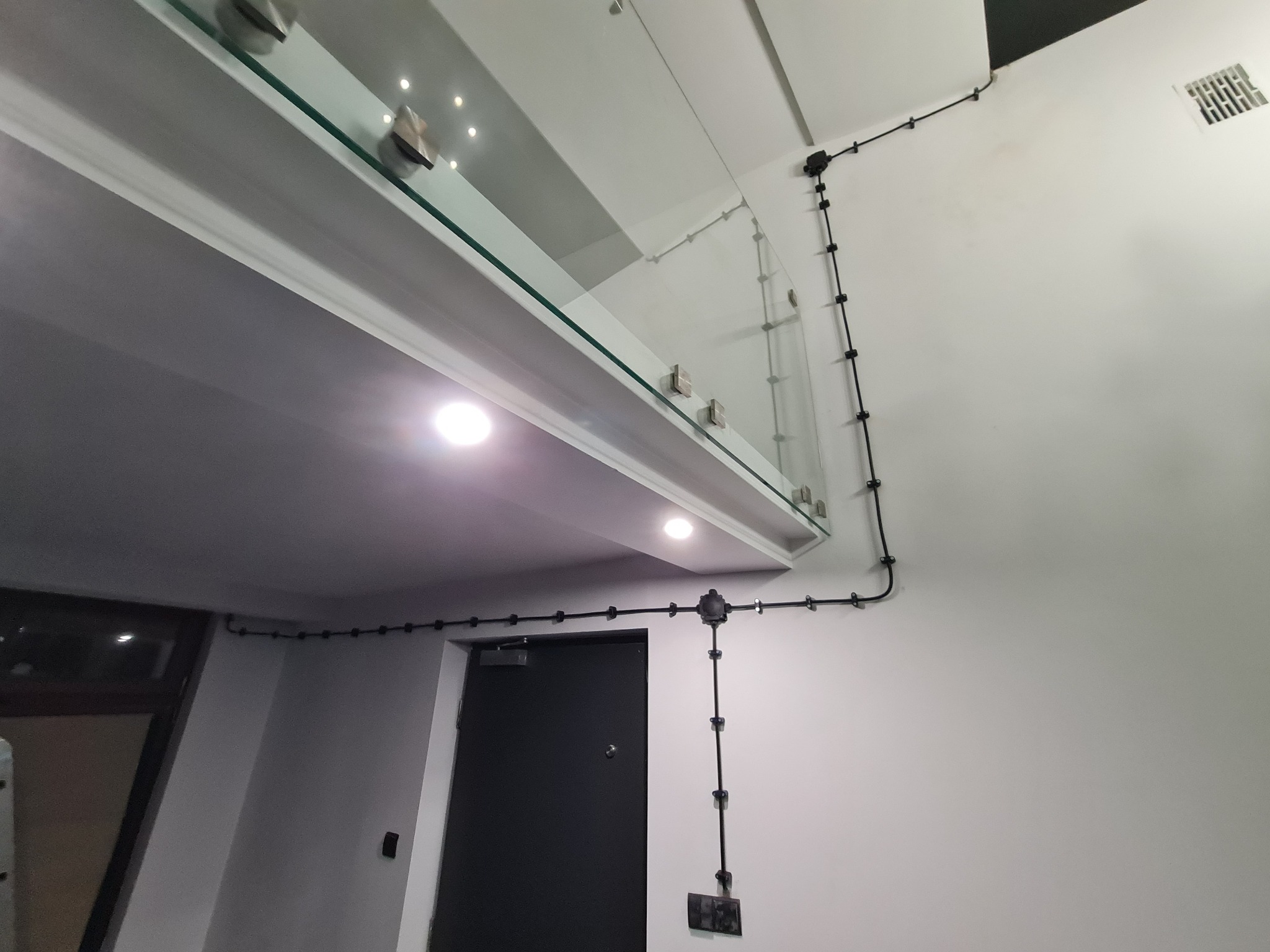 Wnętrze z widoczną instalacją elektryczną natynkową w czarnych rurkach, biegnącą wzdłuż białej ściany i sufitu, obok szklanej balustrady i czarnych drzwi.