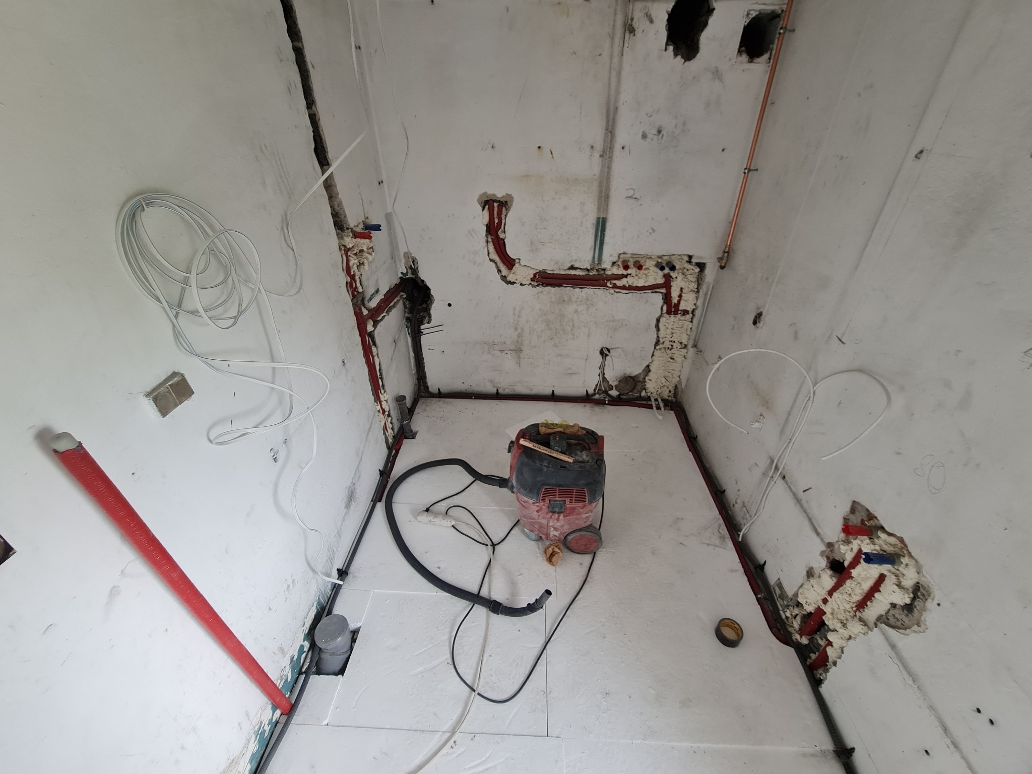 Remont łazienki w trakcie: instalacja rur miedzianych, przewodów elektrycznych, izolacji podłogowej i odkurzacz przemysłowy. Widoczne ślady po kuciu w ścianach.