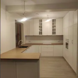 Jasna kuchnia z drewnianym blatem i wyspą, białe fronty szafek z przeszkleniami, minimalistyczne oświetlenie, parkiet w odcieniach beżu, nowoczesny design.