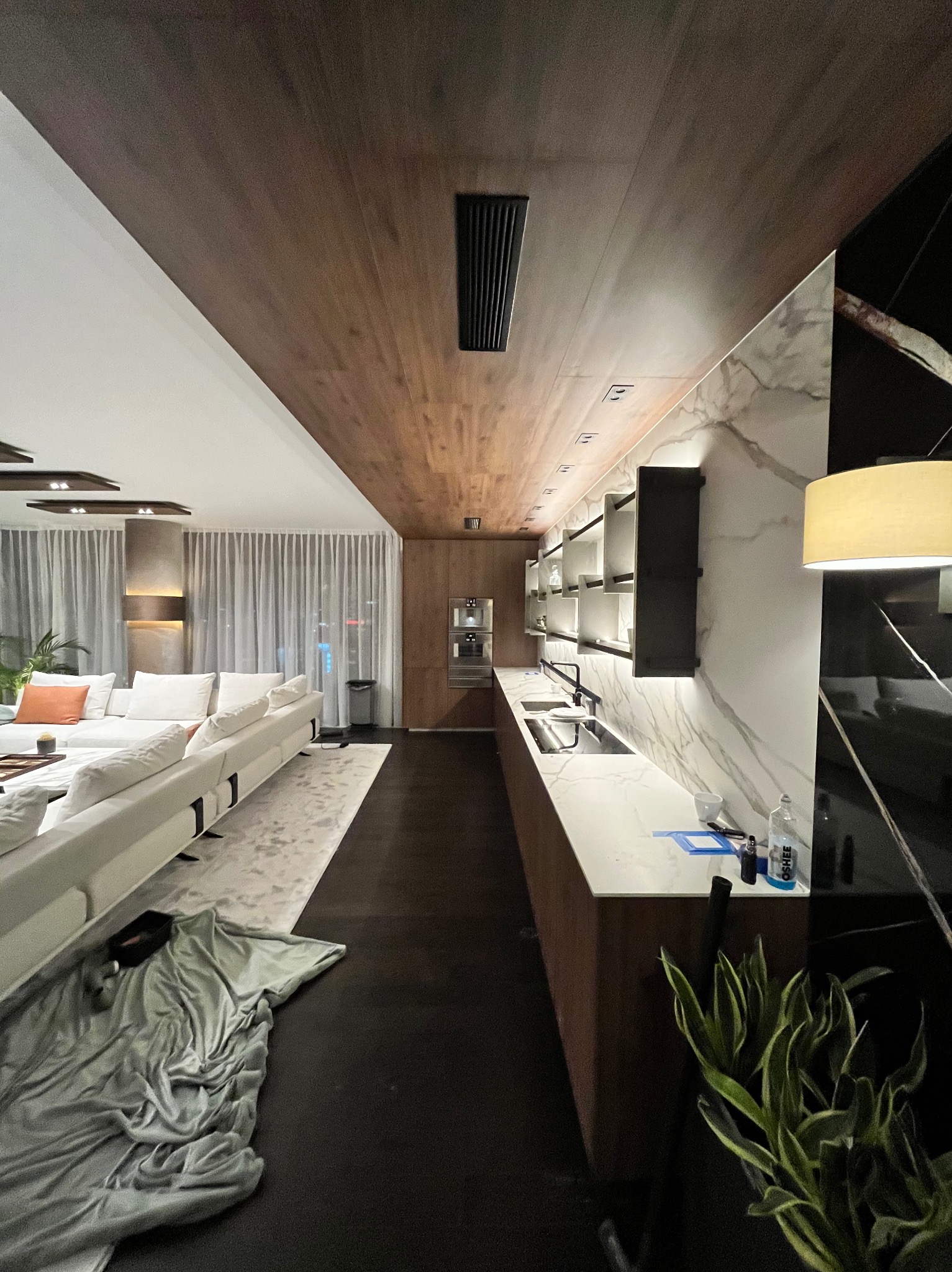 Nowoczesna kuchnia z blatem z białego marmuru i drewnianym sufitem, oświetlona punktowo. Widok na salon z białymi sofami i zasłonami.