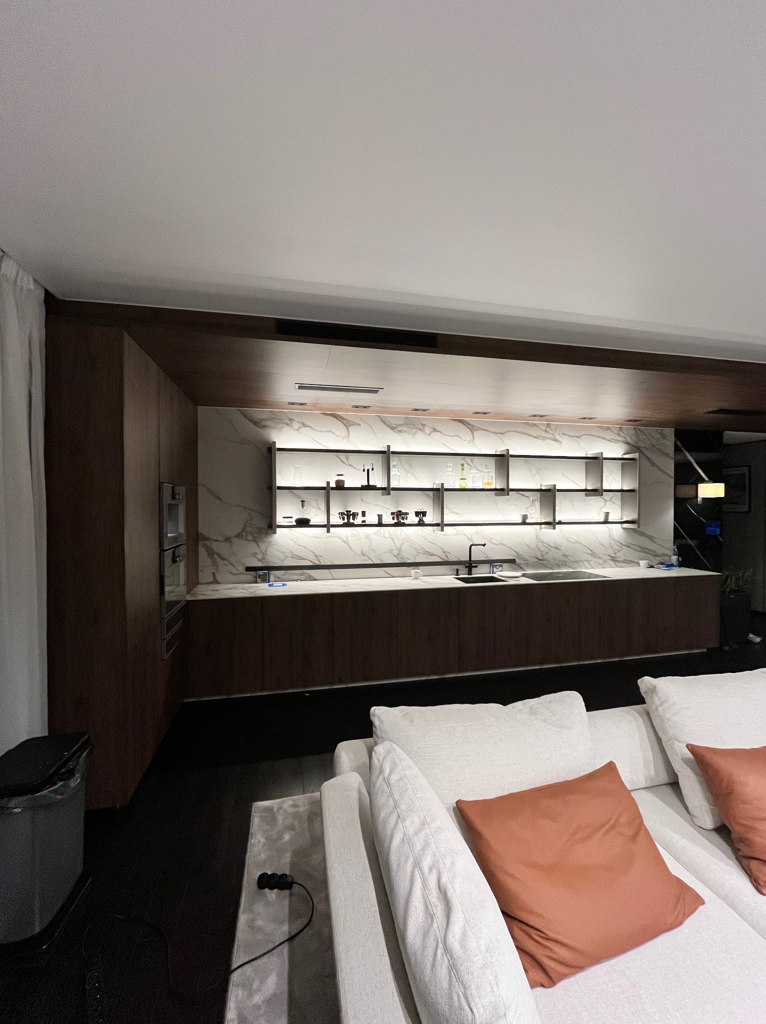 Nowoczesna kuchnia z ciemnego drewna i marmuru, oświetlona półkami, widok z salonu z białą sofą i rdzawymi poduszkami.