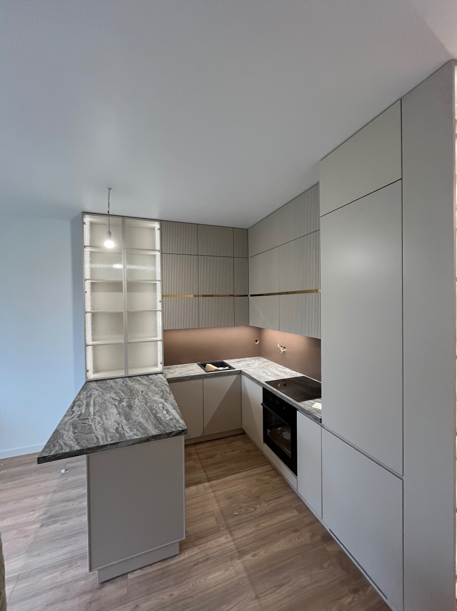 Nowoczesna kuchnia z szarymi frontami i blatem imitującym marmur. Uwagę przyciąga wysoka zabudowa z ryflowanymi elementami i podświetlana witryna.