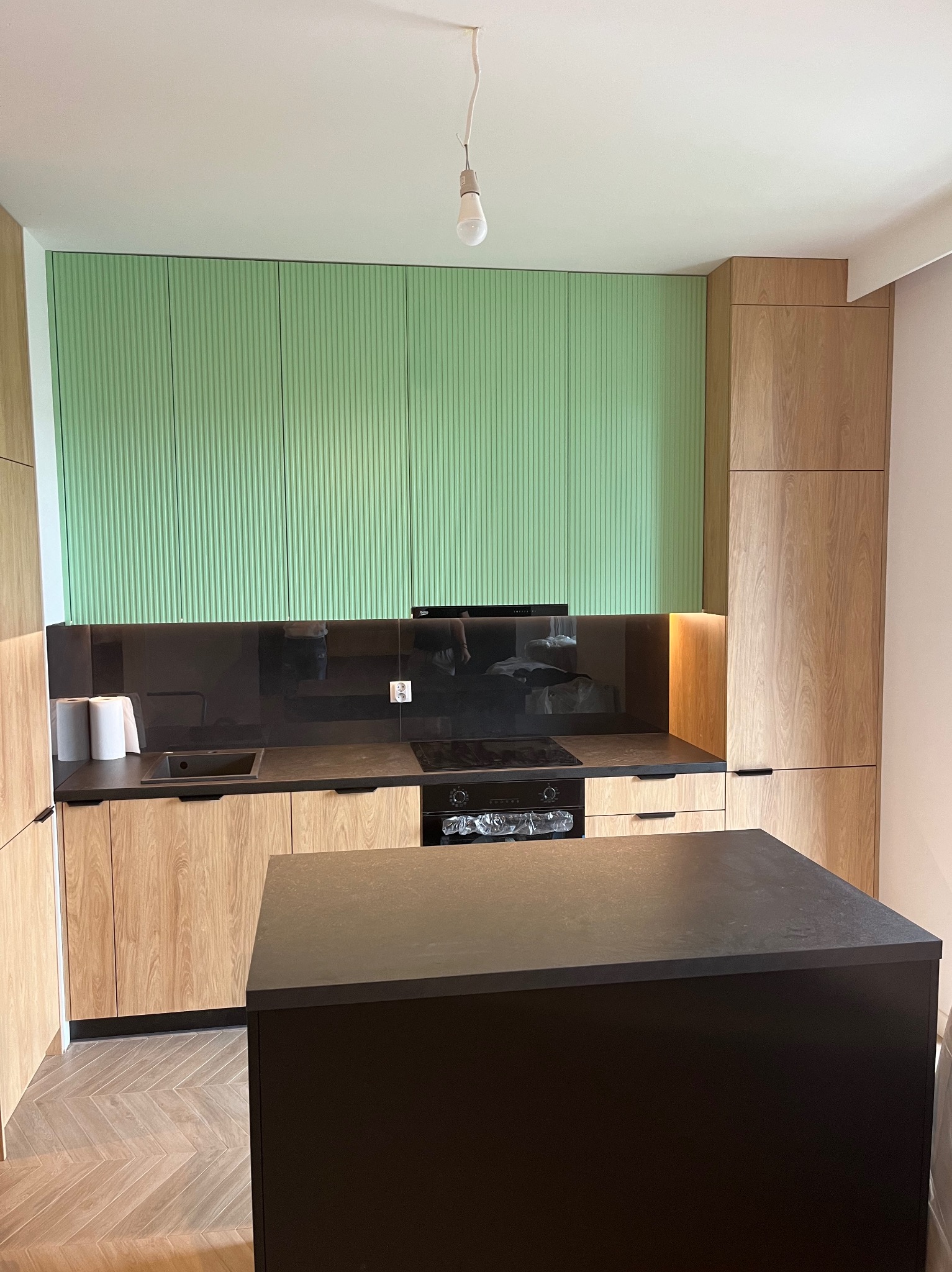 Nowoczesna kuchnia z zielonymi szafkami górnymi o ryflowanej fakturze i wyspą kuchenną na parkiecie w jodełkę. Blat z czarnego kamienia.