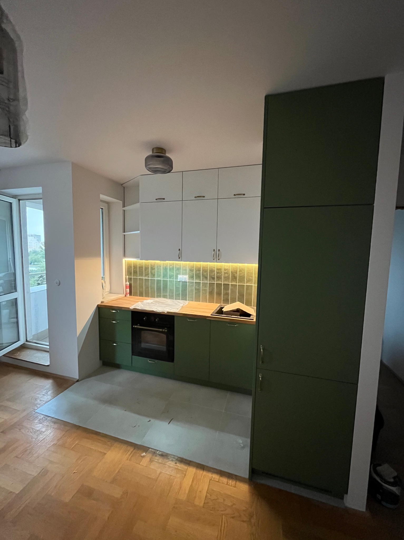 Nowoczesna kuchnia z zielonymi szafkami dolnymi i białymi górnymi, podświetlane płytki, drewniany blat, parkiet. Widok na balkon przez otwarte drzwi.