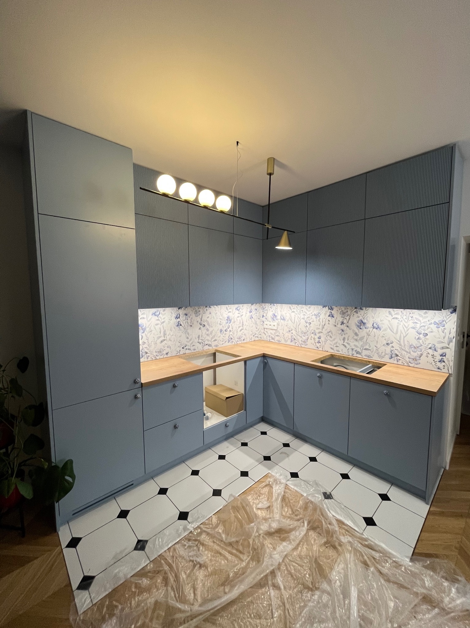 Niebieska kuchnia na wymiar z drewnianym blatem i wzorzystą ścianą nad blatem. Nowoczesne oświetlenie i czarno-biała podłoga.