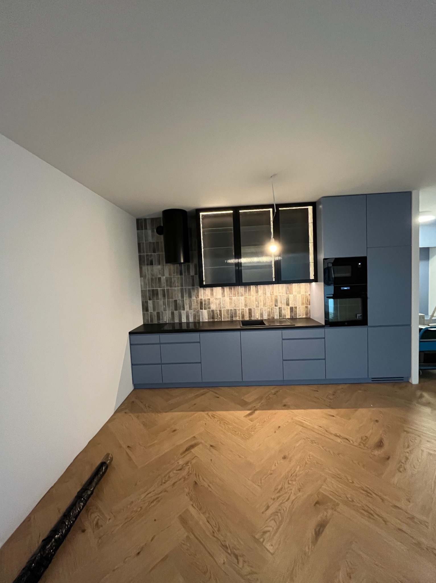 Niebieska zabudowa kuchenna z czarnym okapem i podświetlanymi szafkami wiszącymi, wykończona płytkami imitującymi cegłę. Parkiet w jodełkę na podłodze.