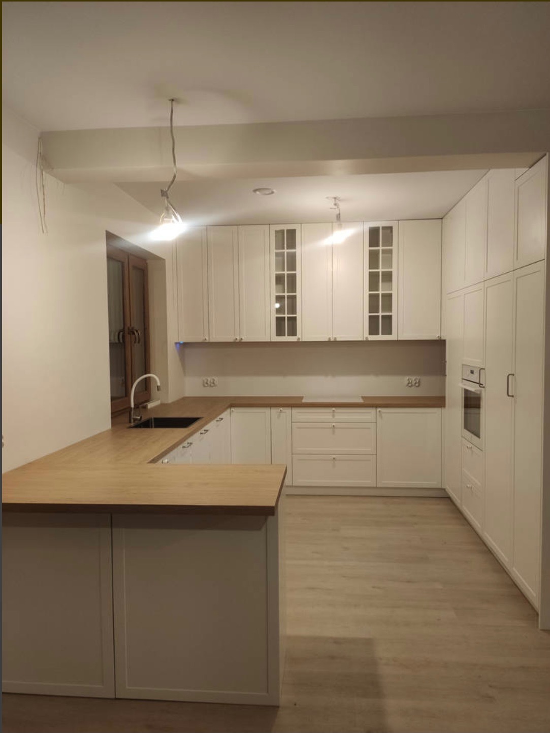 Jasna kuchnia z drewnianym blatem i wyspą, białe fronty szafek z przeszkleniami, minimalistyczne oświetlenie, parkiet w odcieniach beżu, nowoczesny design.