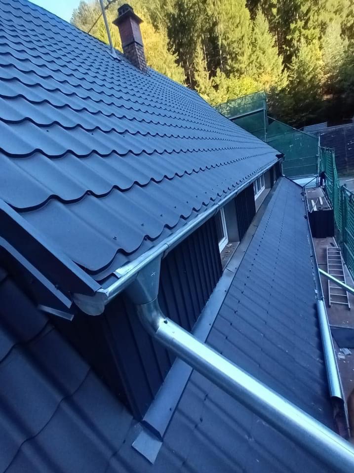 Nowy, ciemny dach z metalowej blachy falistej, z zamontowanymi srebrnymi rynnami; w tle drzewa i komin