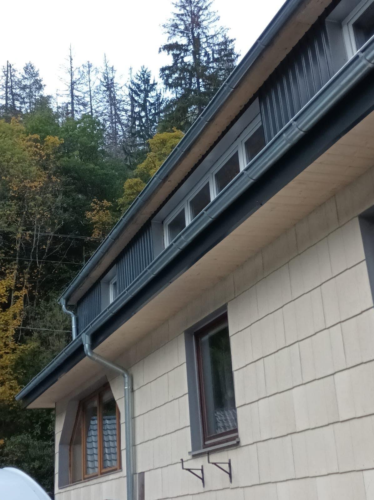 Fragment domu z nową, szarą rynną spustową i szarą blachą na dachu, widoczne okna i elewacja w jasnym kolorze.
