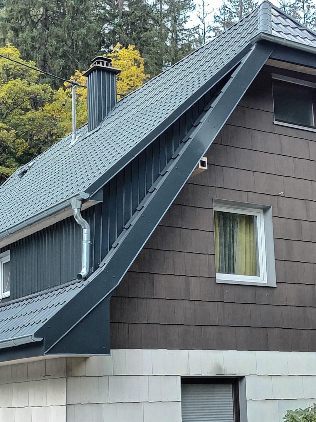 Nowoczesny dom z ciemnoszarą dachówką, widoczne rynny, komin i okna. Detal architektoniczny na styku dachu i ściany z nietypowym oknem.