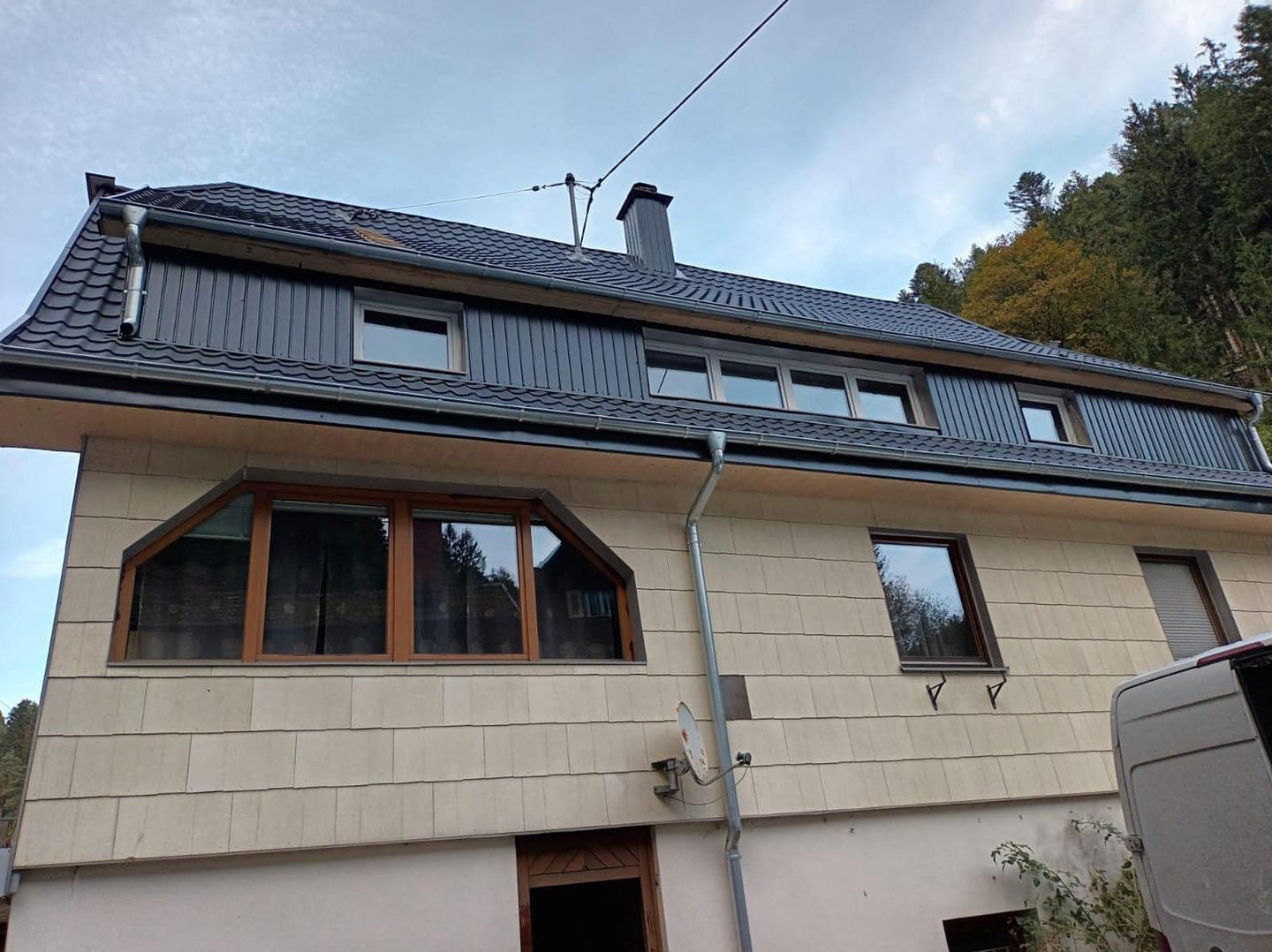 Dom jednorodzinny z nowym, ciemnoszarym dachem z blachodachówki, widoczne rynny i komin, elewacja pokryta jasnymi płytkami imitującymi drewno, okna z drewnianymi ramami.