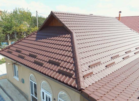 Widok z góry na brązowy dach z dachówki ceramicznej, z widocznymi oknami budynku i fragmentem innego dachu w tle.