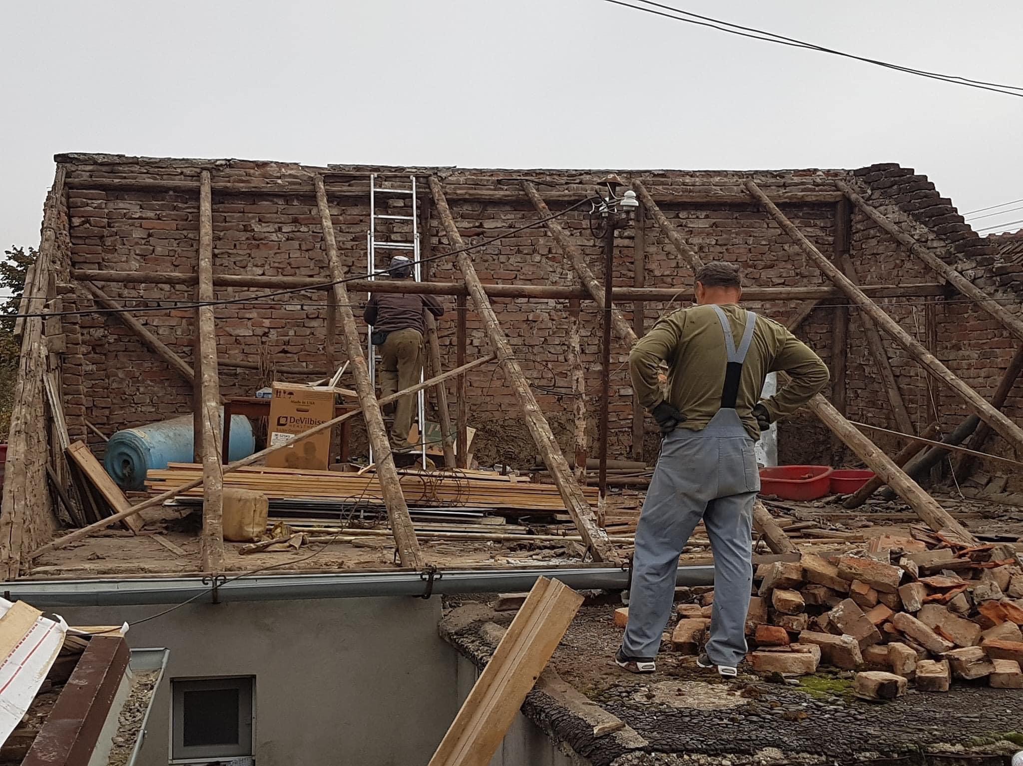 Demontaż starego dachu z cegły, widoczna konstrukcja więźby, jeden pracownik na drabinie, drugi z rękami na biodrach obserwuje postęp prac, sterta gruzu z cegieł na dachu.