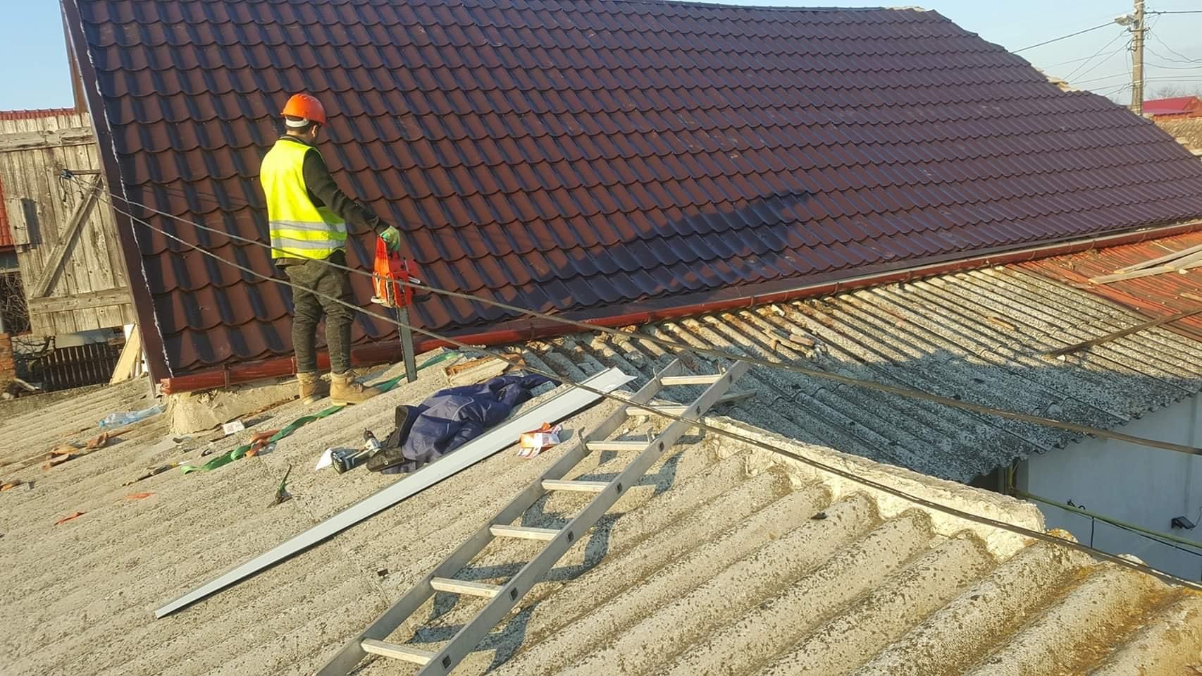 Pracownik w kamizelce i kasku, stojący na dachu z nową, brązową dachówką, obok starego dachu z eternitu i drabiny, z piłą do cięcia dachówek w dłoni.