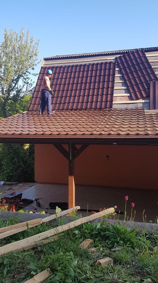 Dekarz w niebieskim kasku montuje brązową blachodachówkę na dachu z widocznymi łatami, częściowo zakończona wymiana pokrycia, widok z dołu na tle zieleni i błękitnego nieba.