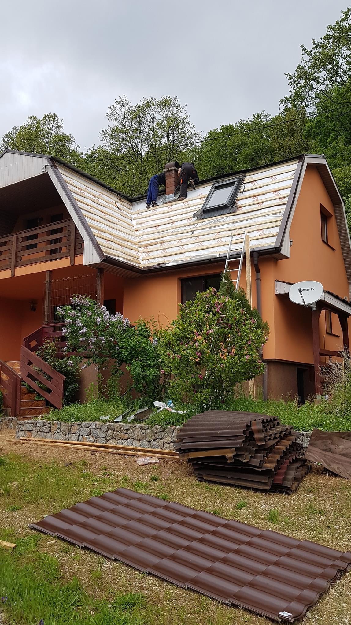 Dwóch dekarzy pracuje na dachu domu jednorodzinnego, widoczne przygotowane brązowe dachówki na trawniku, drabina oparta o dach, okno dachowe i komin.