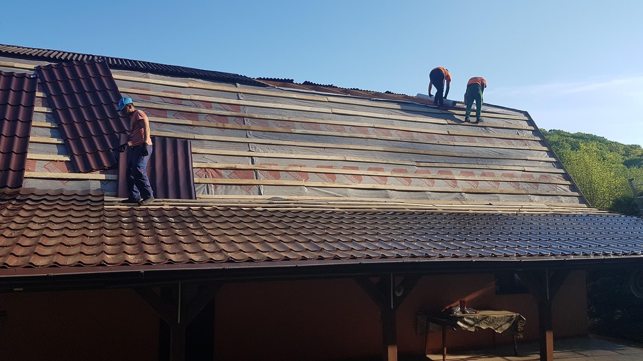 Montaż brązowych paneli blaszanych na dachu domu jednorodzinnego, widoczni dekarze w strojach roboczych na tle błękitnego nieba.