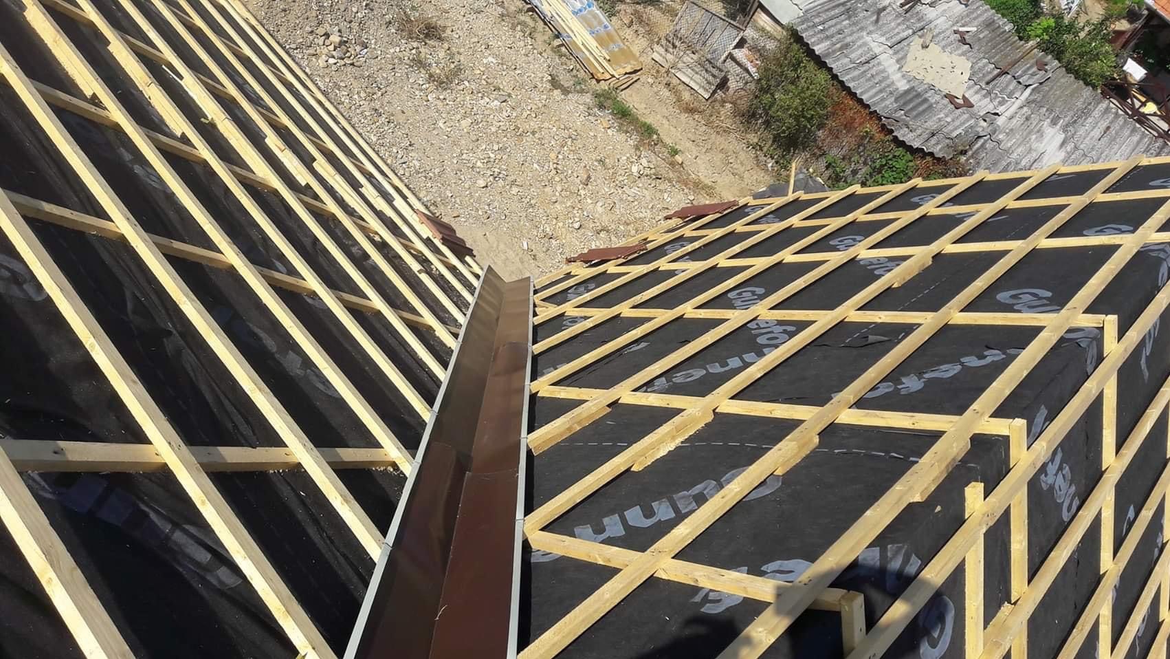 Widok z góry na konstrukcję drewnianą dachu pokrytą czarną membraną, z zamontowaną brązową obróbką blacharską przy krawędzi, w tle fragmenty innych dachów i ziemia.