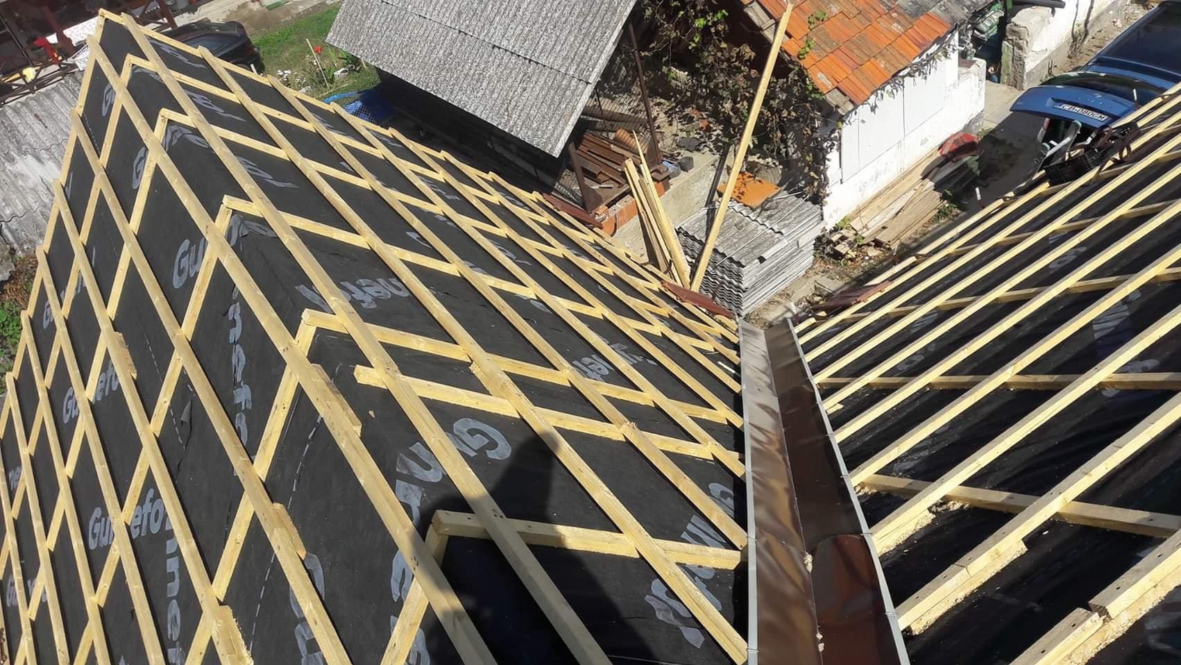 Widok z góry na konstrukcję dachu w trakcie budowy, z widocznymi łatami drewnianymi ułożonymi na czarnej membranie, z fragmentem rynny i otoczeniem budynków w tle.