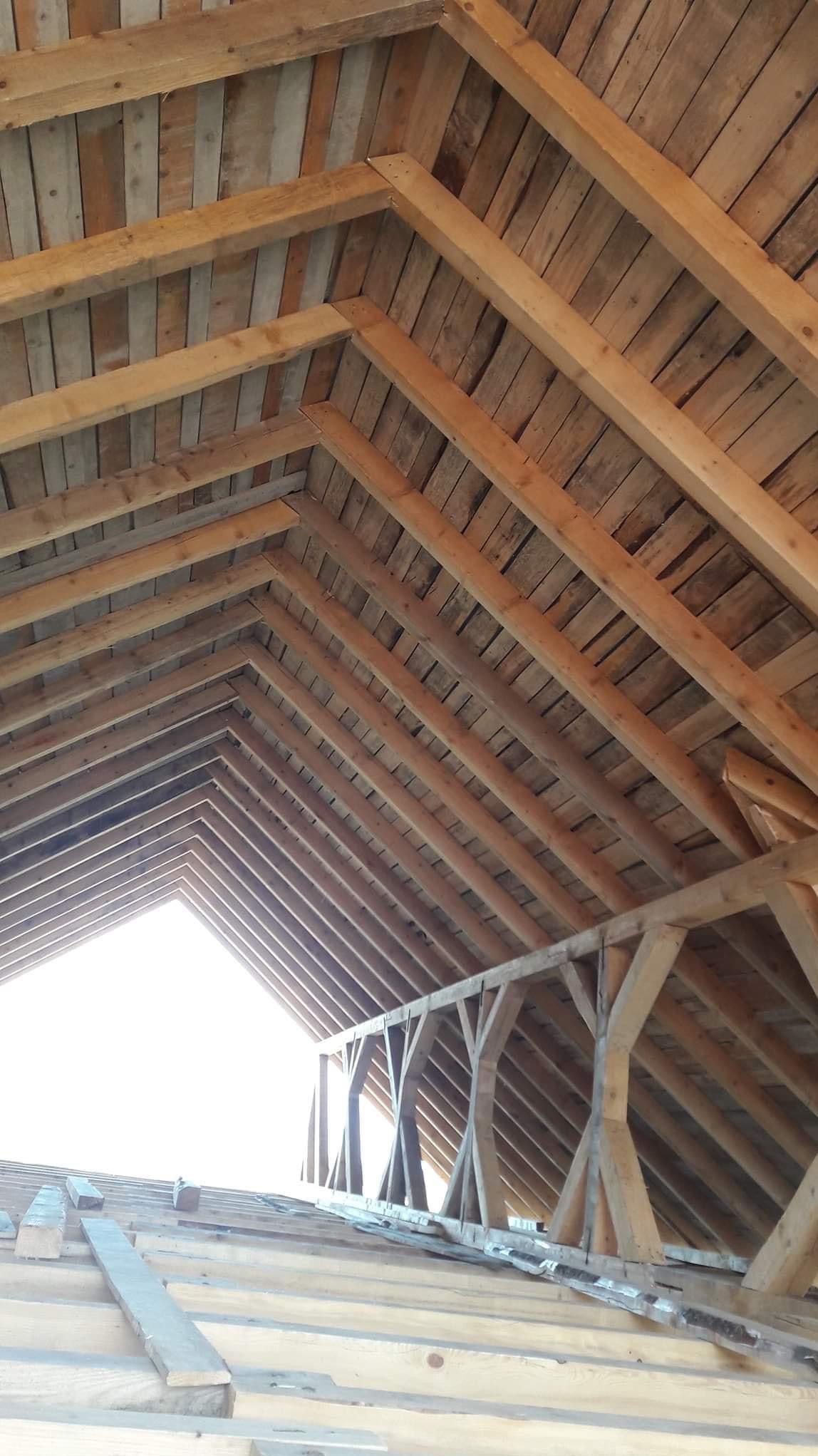 Wnętrze konstrukcji dachu z drewnianymi belkami i deskami, widoczne od wewnątrz, z jasnym prześwitem na końcu.