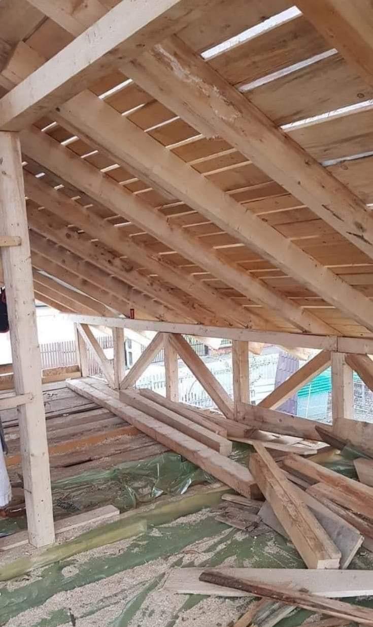Konstrukcja drewniana dachu w trakcie budowy, widoczne krokwie, łaty, kontrłaty i deski, zabezpieczone folią, z widocznymi fragmentami nieociosanych desek na podłodze.
