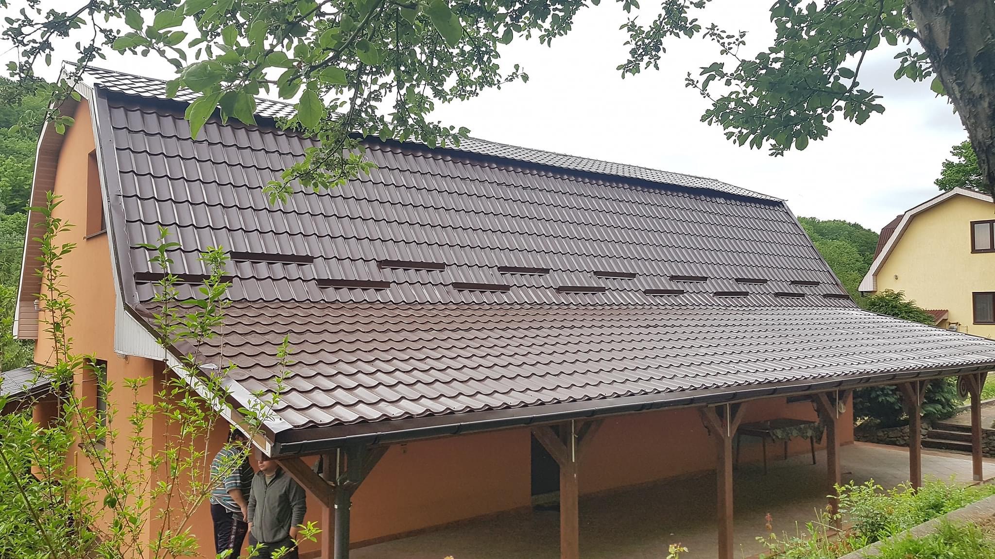 Widok z dołu na brązowy dach z blachodachówki z lukarnami na pomarańczowym domu jednorodzinnym, częściowo przysłonięty gałęziami drzew