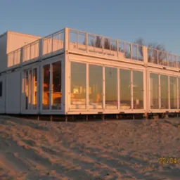 Nowoczesny, dwukondygnacyjny dom z kontenerów morskich, z balkonem na dachu, osadzony na piaszczystym podłożu. Duże przeszklenia odbijają zachodzące słońce.