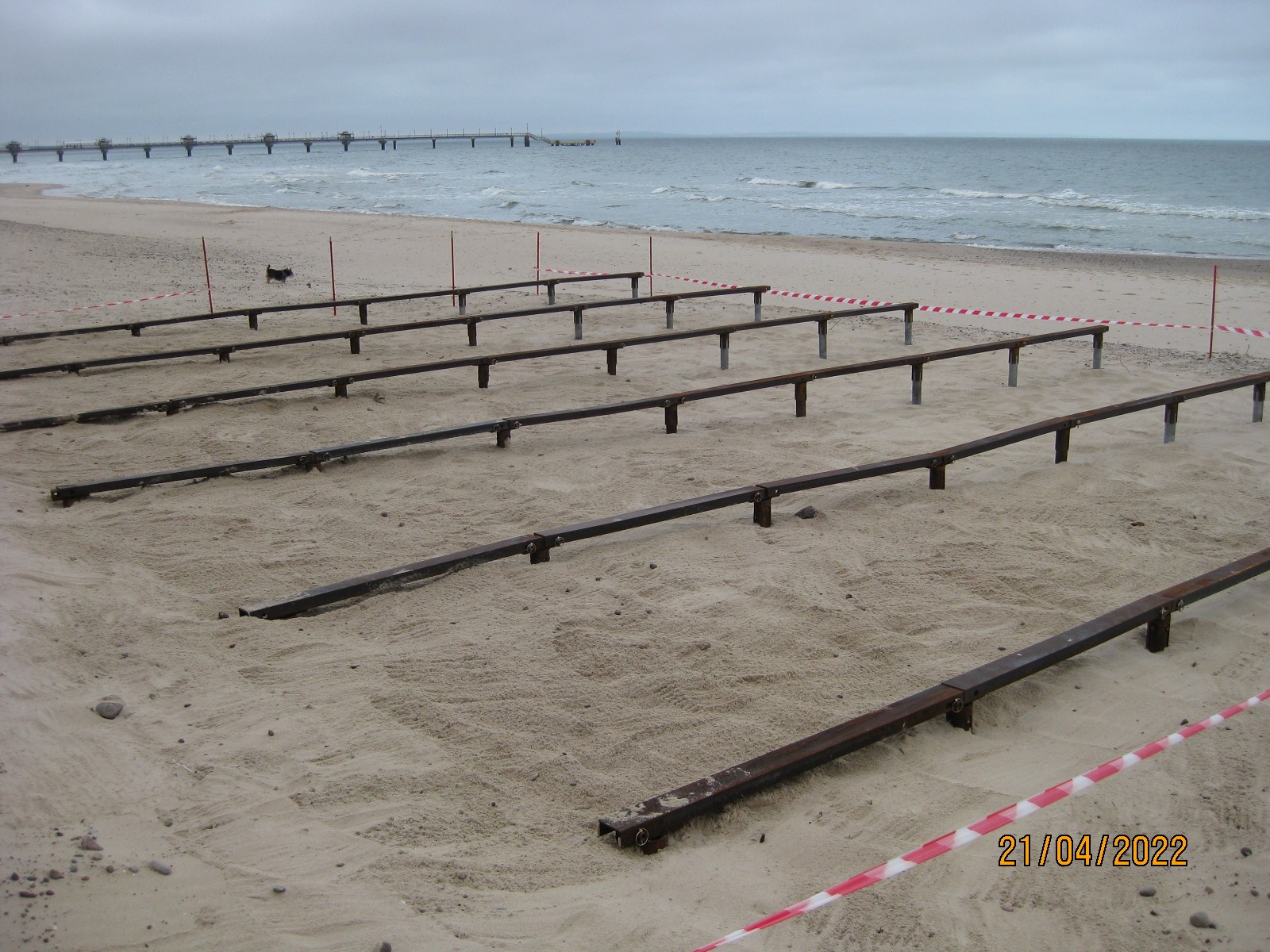 Przygotowanie fundamentów pod konstrukcję na plaży, widoczne metalowe belki i słupy, w tle morze i molo, całość zabezpieczona taśmą ostrzegawczą.