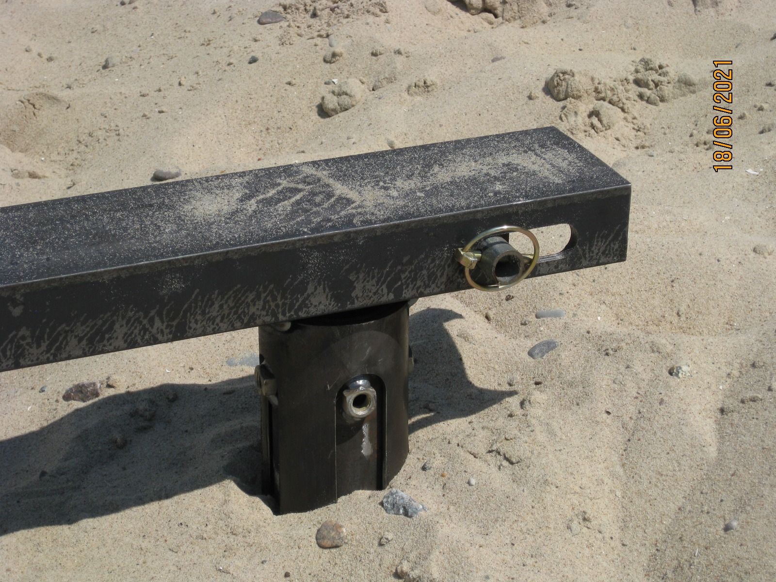 Metalowa konstrukcja wspornika fundamentu, zakotwiczona w piasku, z widocznym sworzniem i śrubami mocującymi. Piasek pokrywa elementy konstrukcji.