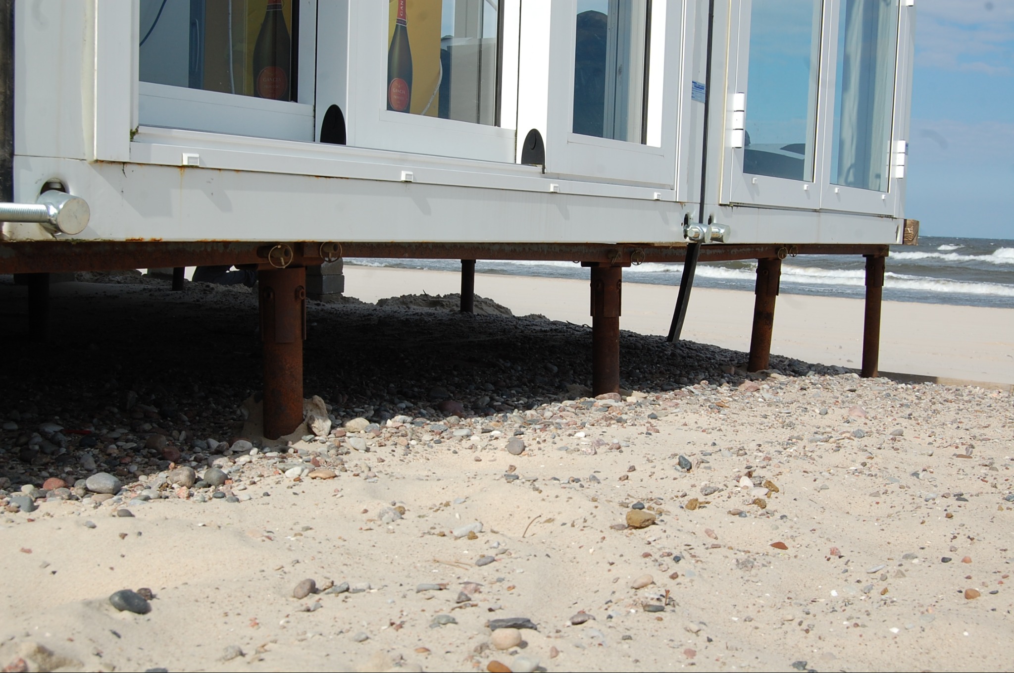 Biały pawilon z oknami, wsparty na metalowych słupach, stoi na piaszczystej plaży nad morzem. Widoczne fale i drobne kamienie na piasku.