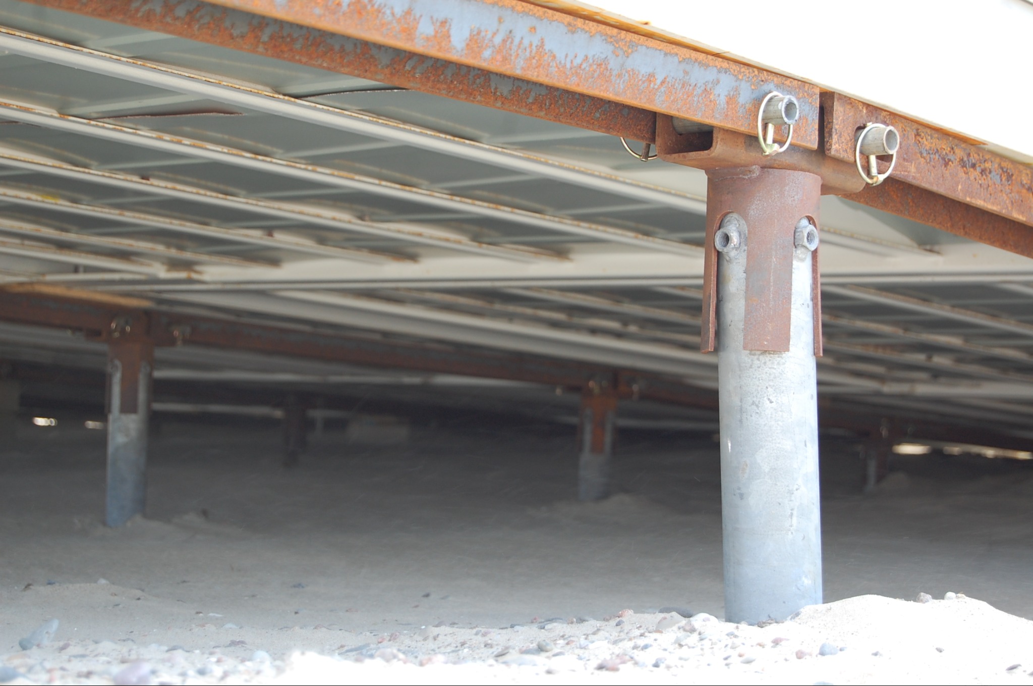 Metalowe podpory konstrukcji na piaszczystym podłożu. Widoczne śruby i elementy mocujące. Konstrukcja zardzewiała w niektórych miejscach. Ujęcie z dołu.